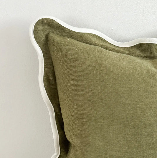 An olive green textured cushion with a white scalloped edge trim, shown against a plain light gray wall.