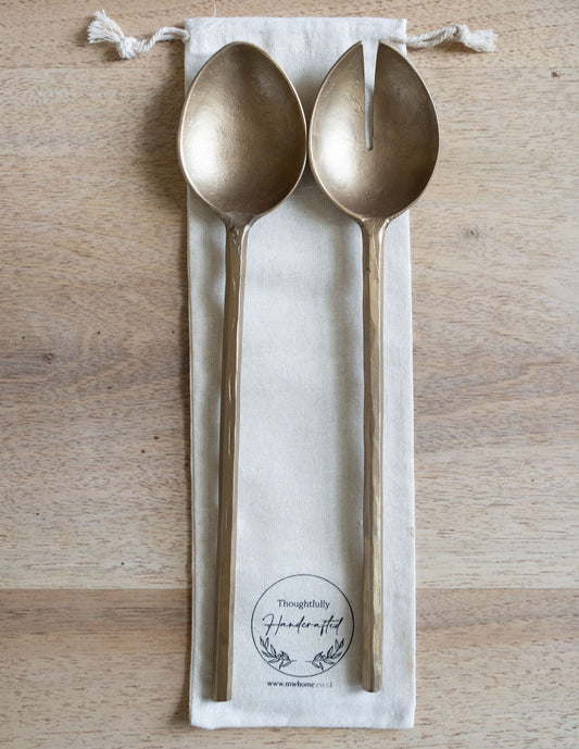 Two brass spoons on a white drawstring bag with a brand logo on a wooden surface