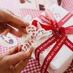 Load image into Gallery viewer, Gift box with red ribbon and decorative tag on a checkered tablecloth

