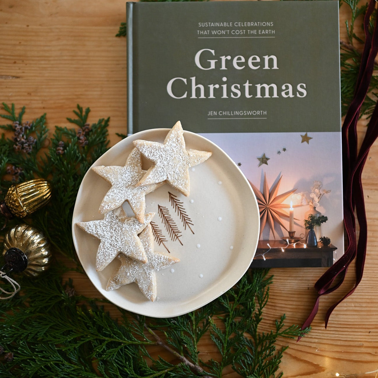 Book titled 'Green Christmas' with star-shaped cookies on a plate, surrounded by greenery and ornaments on a wooden surface.