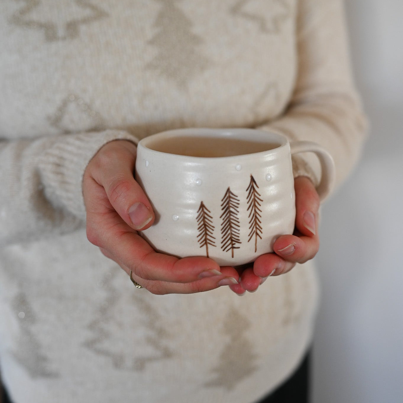 Person holding a white mug with tree design, wearing a beige sweater.