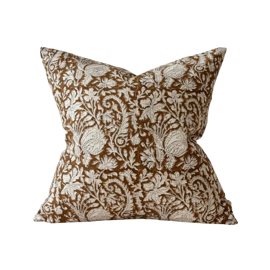A square decorative cushion featuring an intricate floral and botanical damask pattern in brown and cream, set against a plain cream background.