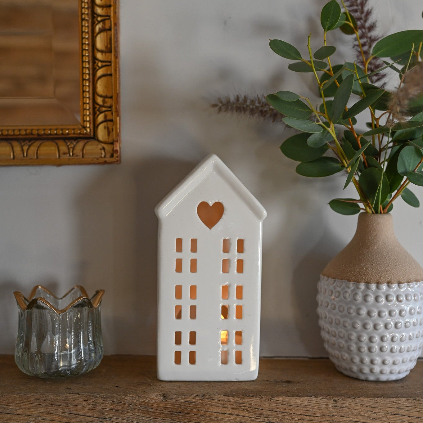 Decorative house-shaped lantern with a heart on a wooden surface, surrounded by plants and a mirror.