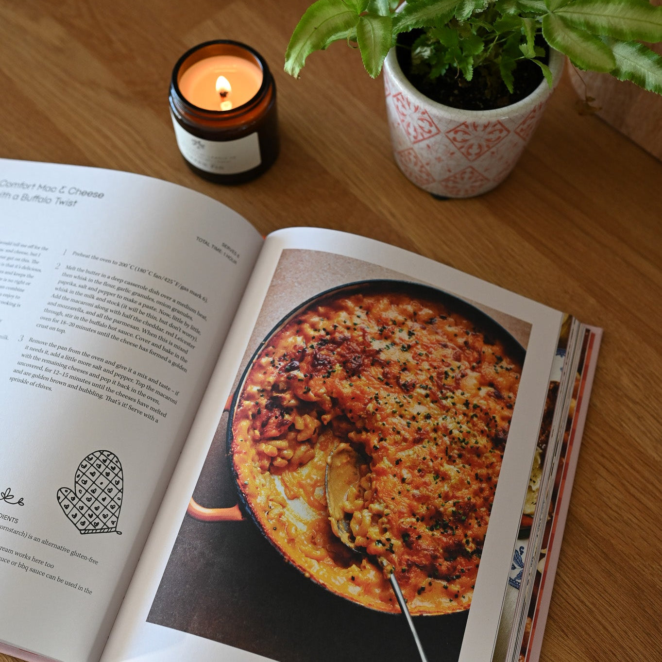 Open cookbook showing a pizza recipe on a wooden table with a lit candle and plant.