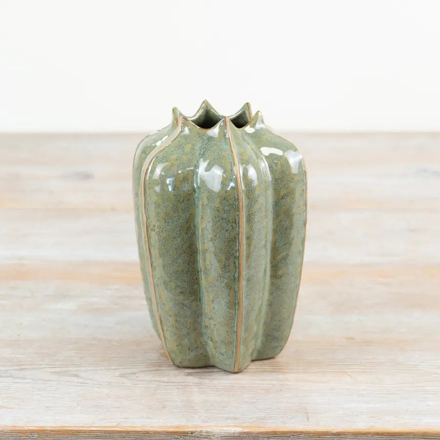 A small ceramic vase with a glossy green glaze and a ribbed, ribbed silhouette, standing on a light wooden surface against a white background.
