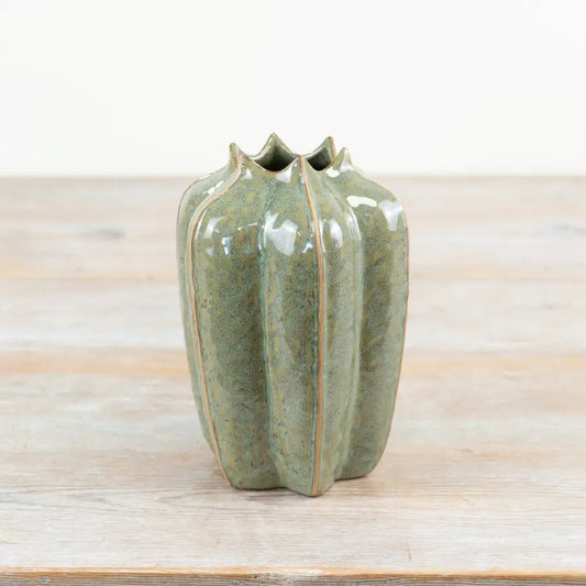 A small ceramic vase with a glossy green glaze and a ribbed, ribbed silhouette, standing on a light wooden surface against a white background.