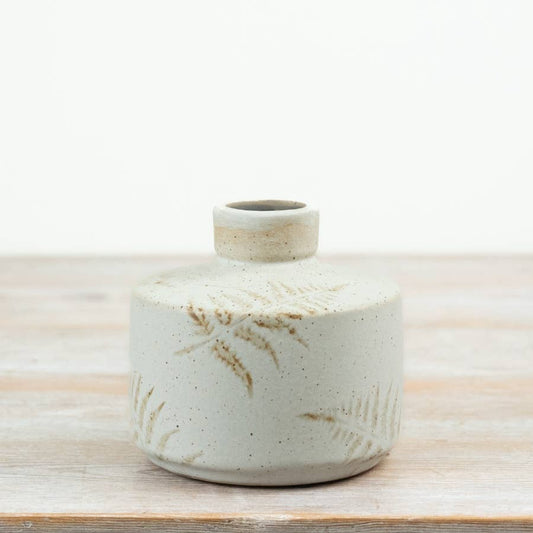 A small round ceramic vase with a short neck, featuring a speckled off-white glaze and embossed leaf patterns, set on a light wooden surface against a white background.