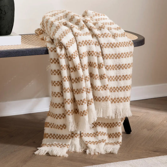 A textured throw blanket with a cream and brown checkerboard pattern and fringed edges, draped over a woven bench in a modern room setting.