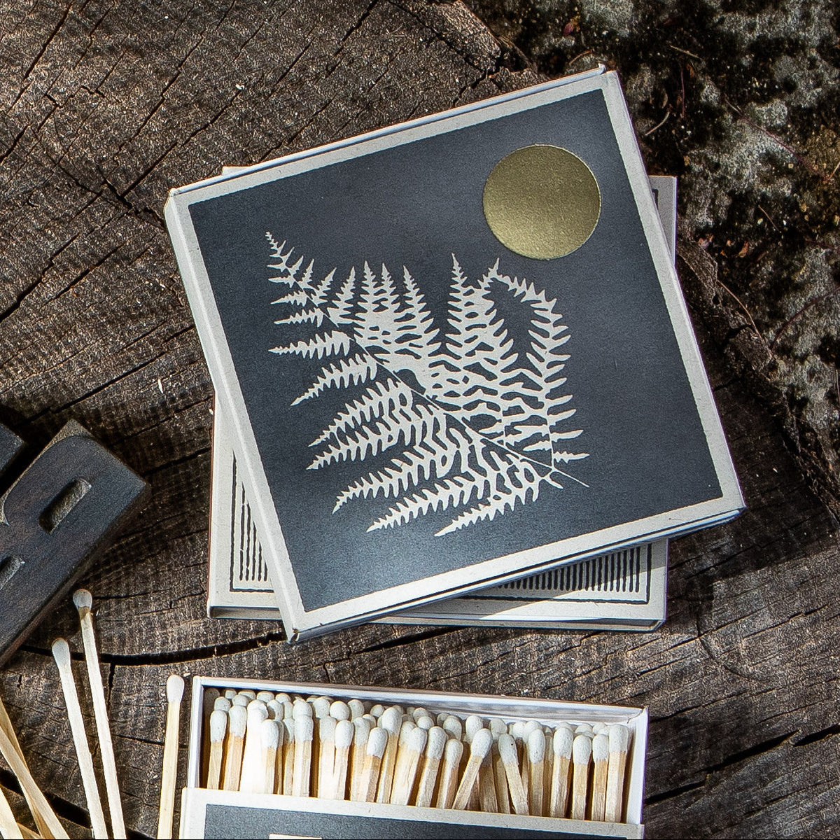 A box of square matchboxes with a dark charcoal cover featuring a white fern leaf design and a gold circular emblem, placed on a rustic wood surface next to an open box of wooden matches.