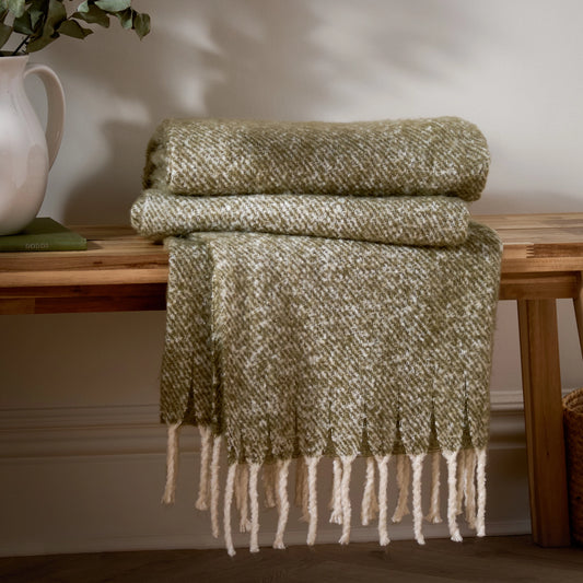 Green woven blanket with fringes draped over a wooden surface.