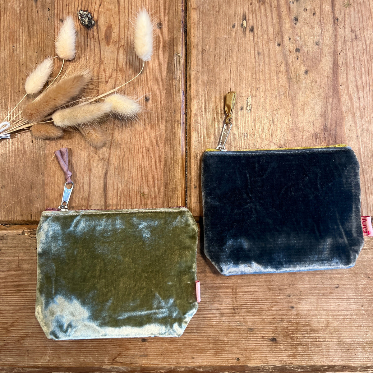 Two velvet pouches, one green and one blue, on a wooden surface with dried flowers.