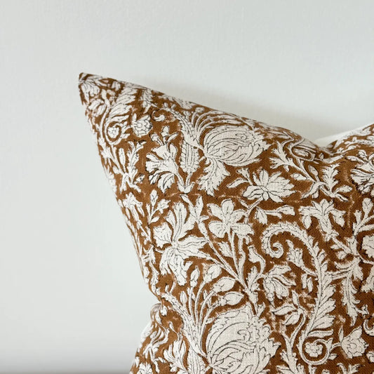 A close-up of a decorative cushion featuring an intricate white floral and botanical pattern woven onto a warm brown velvet background, set against a plain off-white wall.