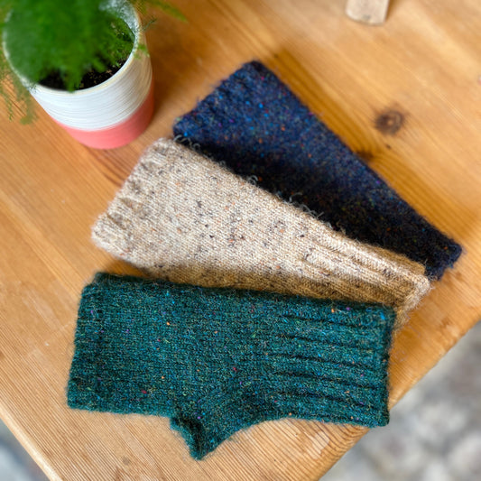 Three knitted ribbed fingerless gloves in forest green, oatmeal beige, and navy blue, arranged diagonally on a light wood tabletop next to a small potted plant.