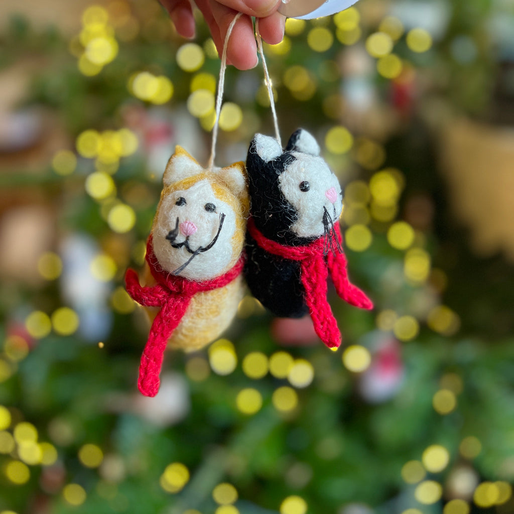 Two small felt cat decorations with scarves held against a blurred Christmas tree background.