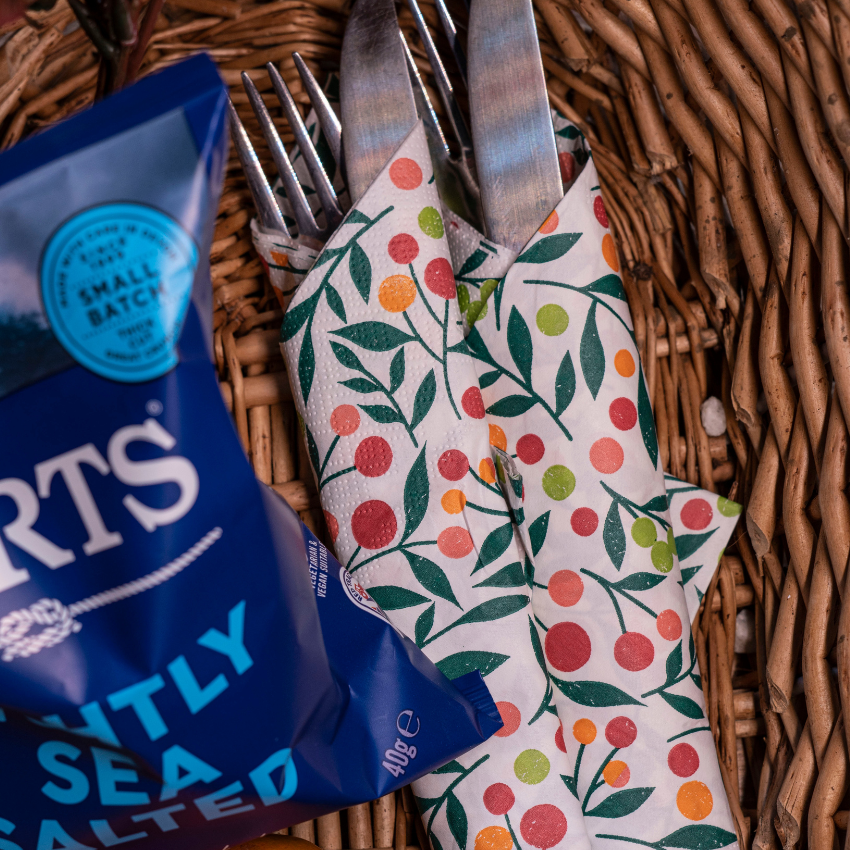 A close-up flat lay of a blue bag of Burts Lightly Sea Salt next to a white napkin with a red, orange, and green fruit pattern, holding silver cutlery, all arranged inside a wicker basket.