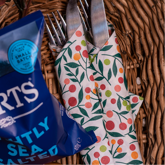 A close-up flat lay of a blue bag of Burts Lightly Sea Salt next to a white napkin with a red, orange, and green fruit pattern, holding silver cutlery, all arranged inside a wicker basket.