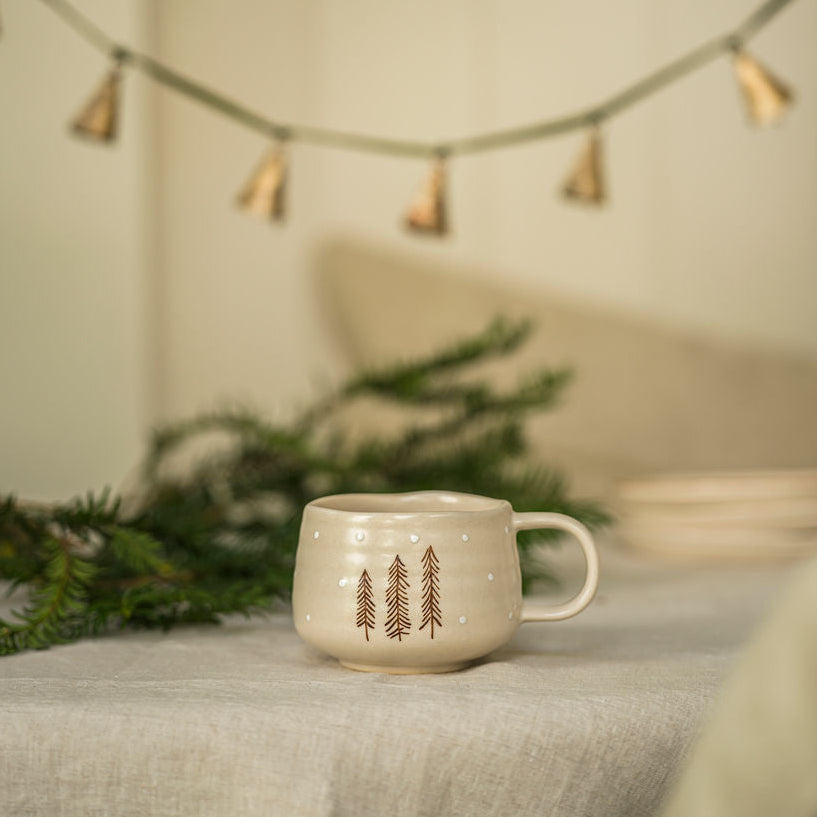 Mug with tree design on a surface with decorative lights and greenery in the background