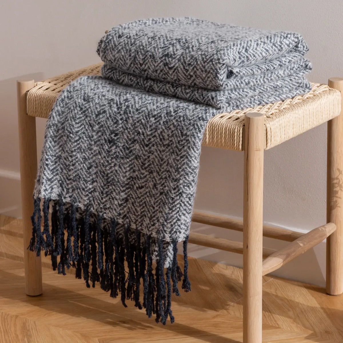A folded and draped throw blanket with a navy and white herringbone pattern and dark navy fringe, displayed on a light wood woven stool against a neutral beige wall.