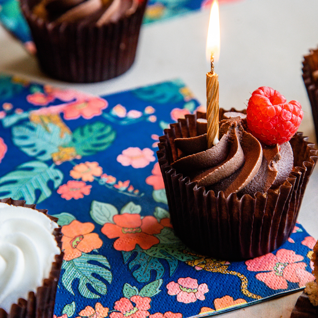 A chocolate cupcake in a brown paper liner with piped chocolate frosting, topped with a single lit candle and a fresh raspberry, resting on a colorful floral napkin.