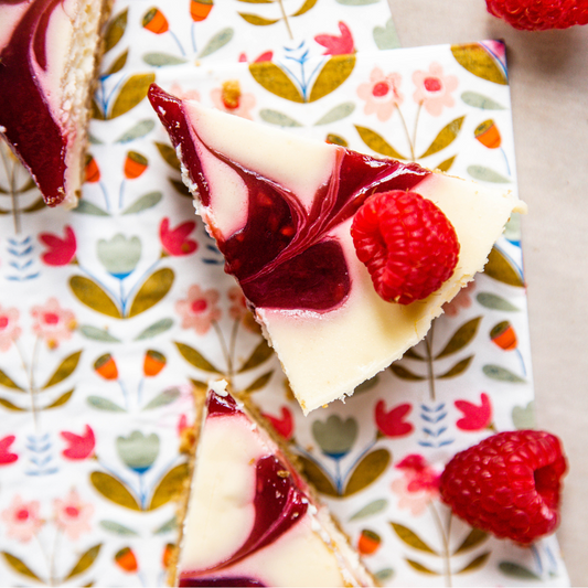A close-up of a triangular slice of cheesecake with a deep berry swirl and a fresh raspberry on top, placed on a floral-patterned napkin with more pie slices and additional raspberries around it.