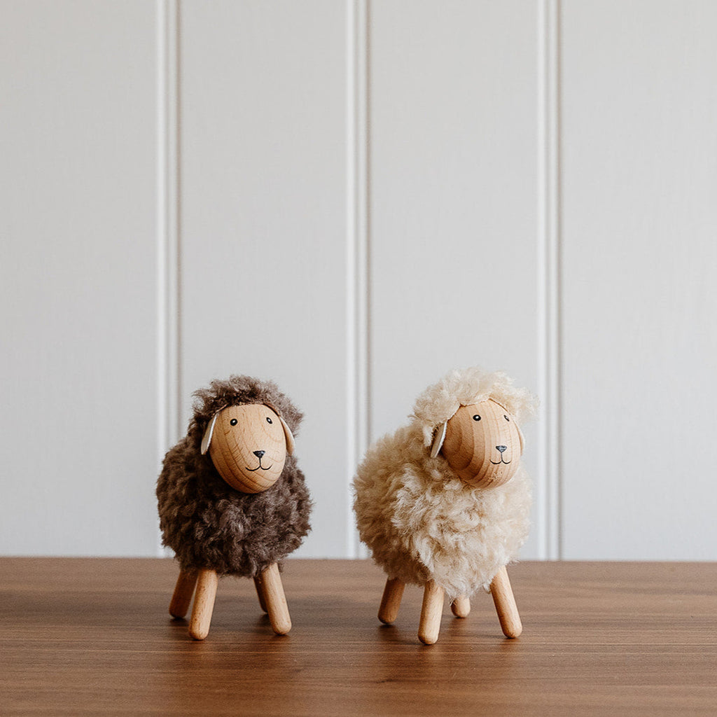 Two wooden sheep, one grey and one cream, standing on a wooden surface with a light gray wall in the background. 