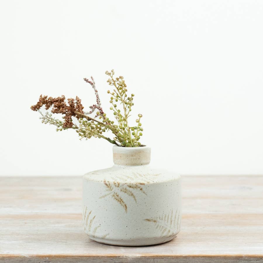 A small round ceramic vase with a short neck, featuring a speckled off-white glaze and subtle gold leaf patterns, holding dried botanical stems on a light wood surface against a white background.