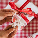 Load image into Gallery viewer, Gift wrapped in red and white paper with a red ribbon on a checkered tablecloth.
